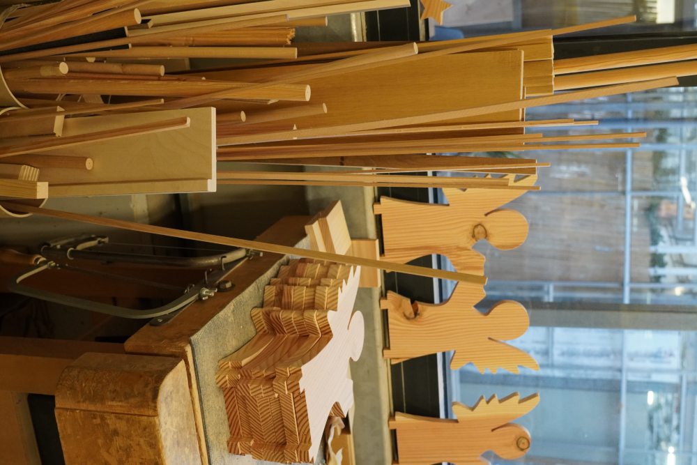 Werkstatt mit Holzbrettern und Holzstangen. Auf einem Tisch liegen ausgesägte Holzformen von Engeln. Im Hintergrund ist ein Fenster mit Gitter zu sehen.