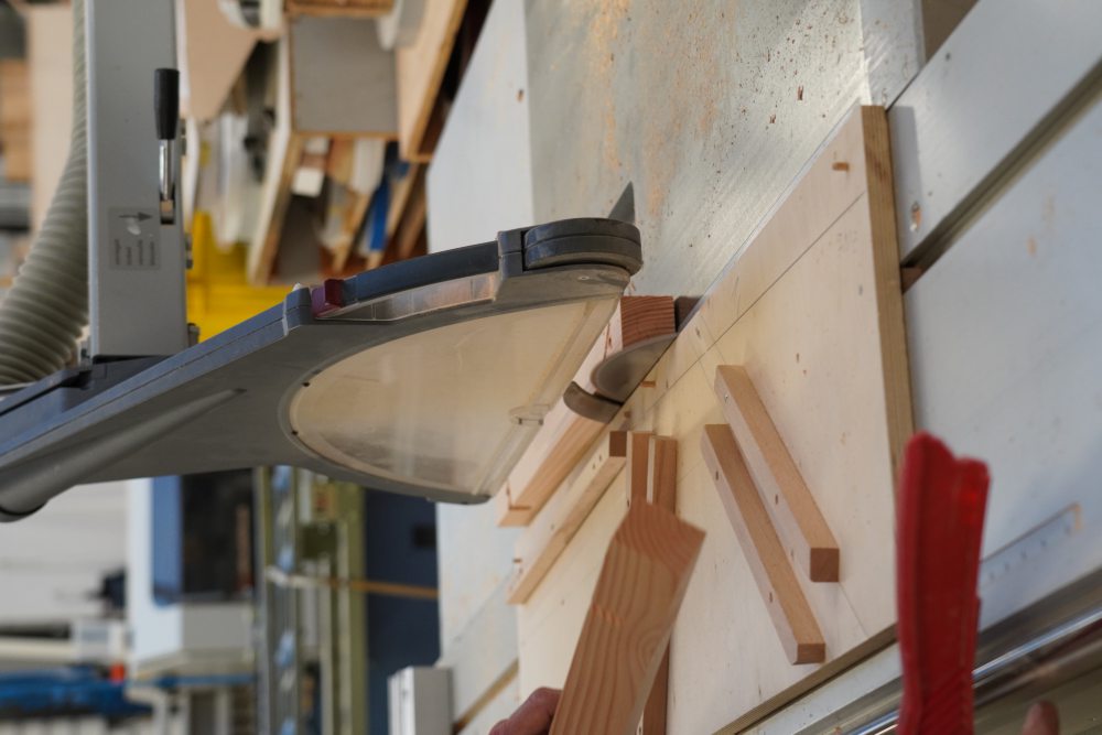 Eine Tischkreissäge schneidet Holzstücke in einer Werkstatt. Die Sägeblattabdeckung schützt das laufende Sägeblatt. Im Hintergrund sind weitere Werkzeuge und Maschinen erkennbar.