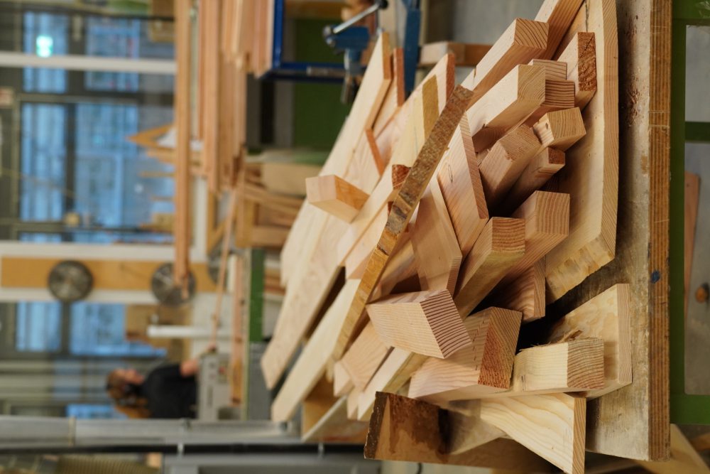 Ein Stapel Holzlatten liegt auf einem Werkstatttisch in einer Schreinerei, im Hintergrund sind Maschinen und eine Person unscharf zu erkennen.
