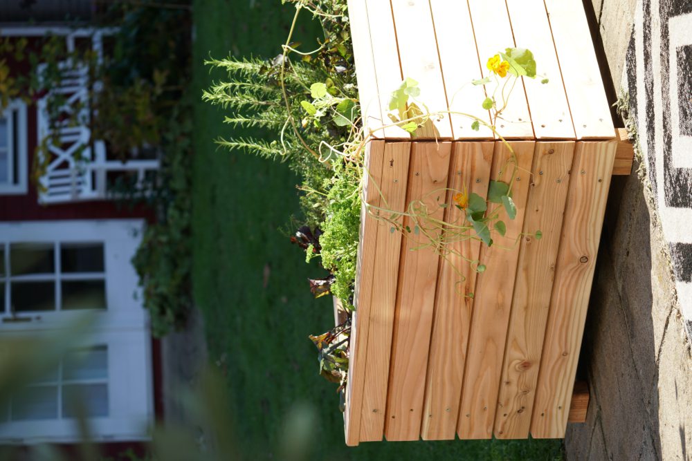 Ein hölzernes Hochbeet steht auf einer Terrasse. Im Hochbeet wachsen verschiedene grüne Pflanzen und Kräuter. Im Hintergrund sind ein Rasen und teilweise ein rotes Gebäude mit weißen Fenstern und Türen zu sehen.  Bild verlinkt zum Produkt