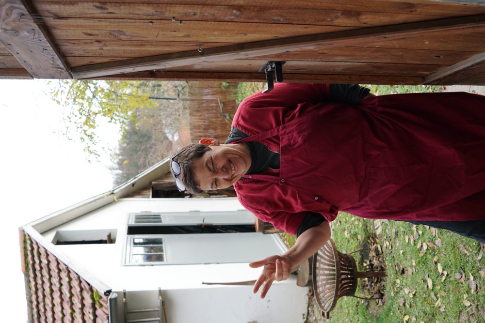 Eine Person in roter Schürze steht lächelnd in einer Türöffnung, zeigt ein Peace-Zeichen, mit einem Garten und einem weißen Gebäude im Hintergrund.
