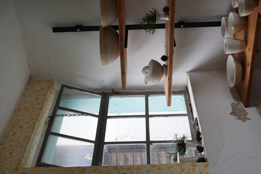 Regal mit Keramikschalen und Pflanzen neben einem Fenster in einem hellen Raum mit Rissen an der Wand. Hölzerne Regalböden sind an einer Metallhalterung befestigt.
