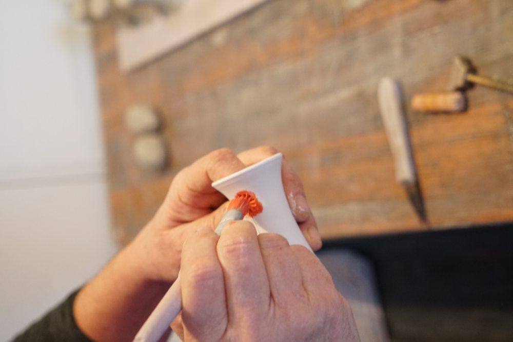 Zwei Hände bemalen eine kleine weiße Keramikvase mit einem Pinsel rot auf einem Holztisch. Im Hintergrund liegen weitere Malutensilien und Werkzeuge.