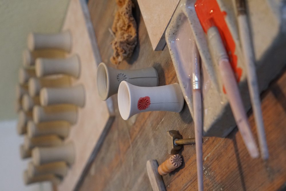 Töpferwerkstatt mit Tonvasen in verschiedenen Stadien der Fertigstellung, Pinseln und Farbpalette im Vordergrund, mehrere unfertige Tonvasen stehen auf einem Regal im Hintergrund.