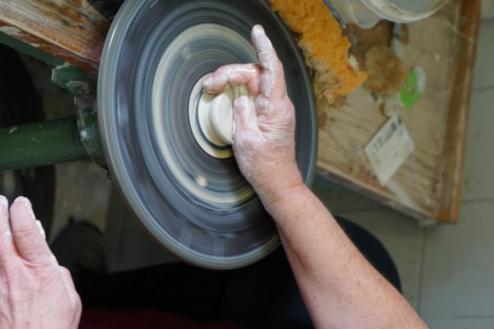 Eine Hand formt Ton auf einer sich drehenden Töpferscheibe. Der Hintergrund zeigt Holztisch mit Werkzeugen und eine weitere Hand stützt sich am Tisch ab.