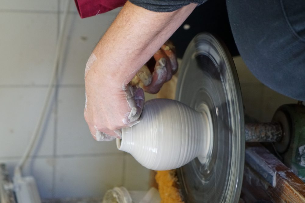 Hände formen eine weiße Tonvase auf einer rotierenden Töpferscheibe in einer Werkstattumgebung.