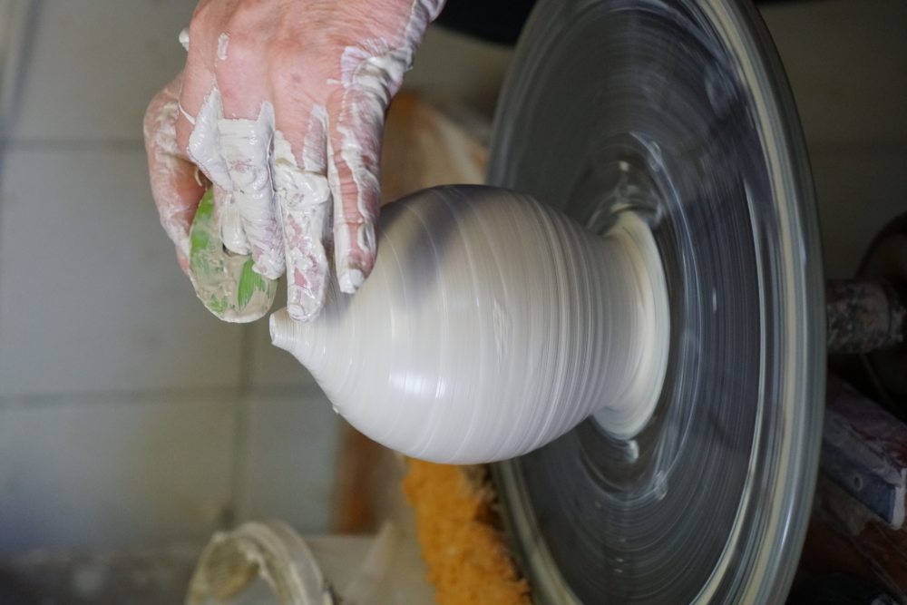 Eine Hand formt eine weiße Tonvase auf einer rotierenden Töpferscheibe; der Raum ist eine Keramikwerkstatt.