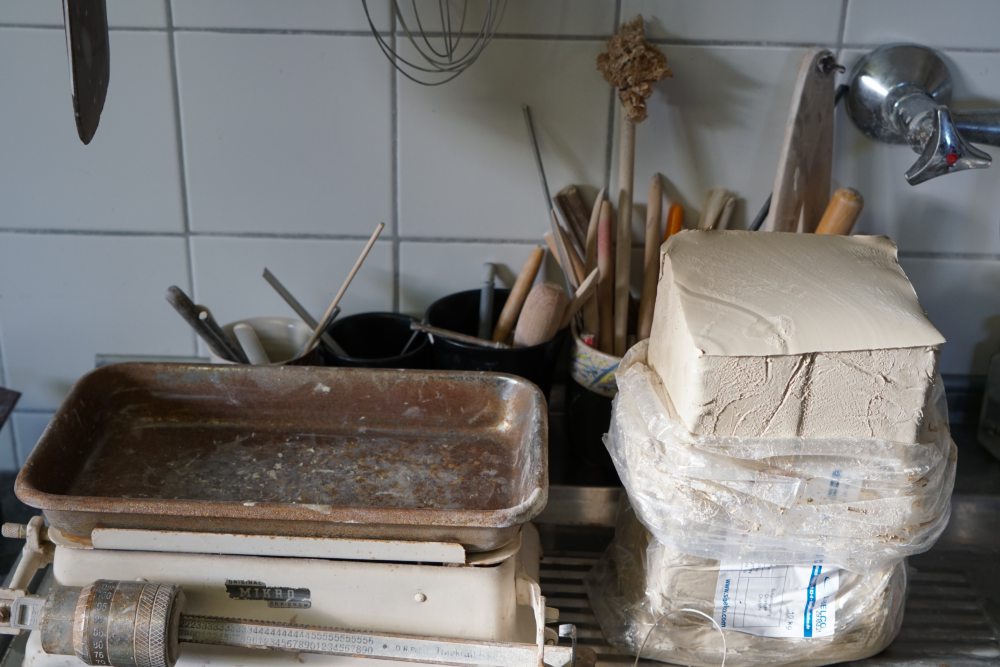 Töpferwerkstatt mit Werkzeugen, Waage und ungebranntem Tonblock auf Metalltisch vor gefliester Wand. Werkzeuge stehen in Behältern; Ton ist in Plastik verpackt.