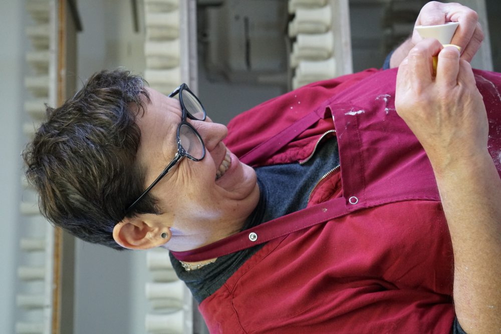Eine Person mit Brille und kurz geschnittenem Haar formt Ton, während sie in einer Werkstatt vor einem Regal mit Keramikprodukten steht. Sie trägt ein rotes Oberteil und eine Schürze.