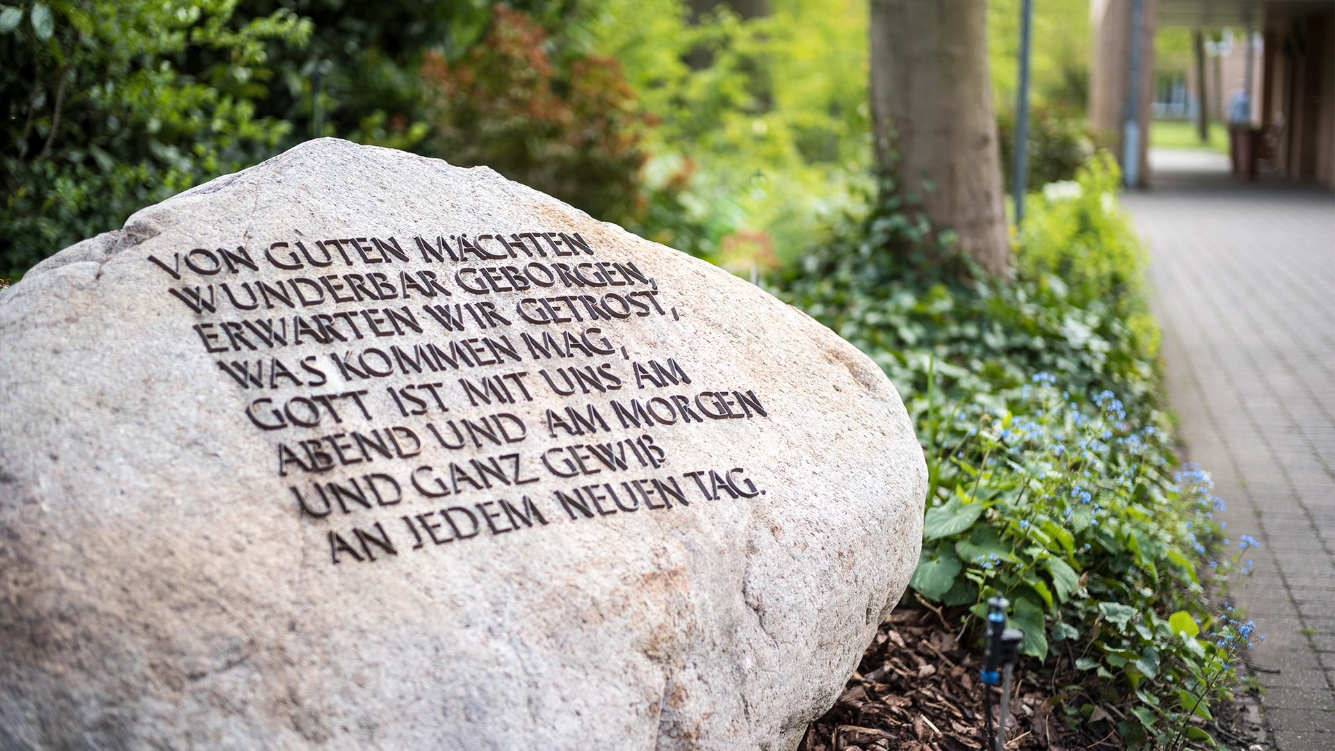 Ein großer Stein mit eingraviertem Text steht in einem Garten. Der Text lautet: Von guten Mächten wunderbar geborgen, erwarten wir getrost, was kommen mag. Gott ist mit uns am Abend und am Morgen und ganz gewiss an jedem neuen Tag.