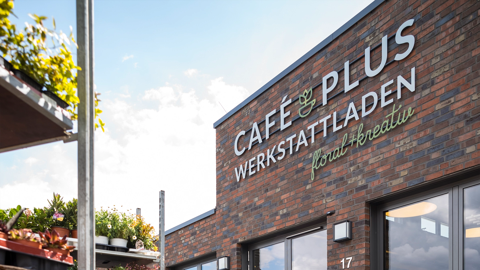 Ziegelfassade mit der Aufschrift „Café & Plus Werkstattladen floral+ kreativ“. Links im Bild stehen Pflanzen auf einem Regal; glasierte Fenster sind unten im Gebäude sichtbar. Der Himmel ist leicht bewölkt.