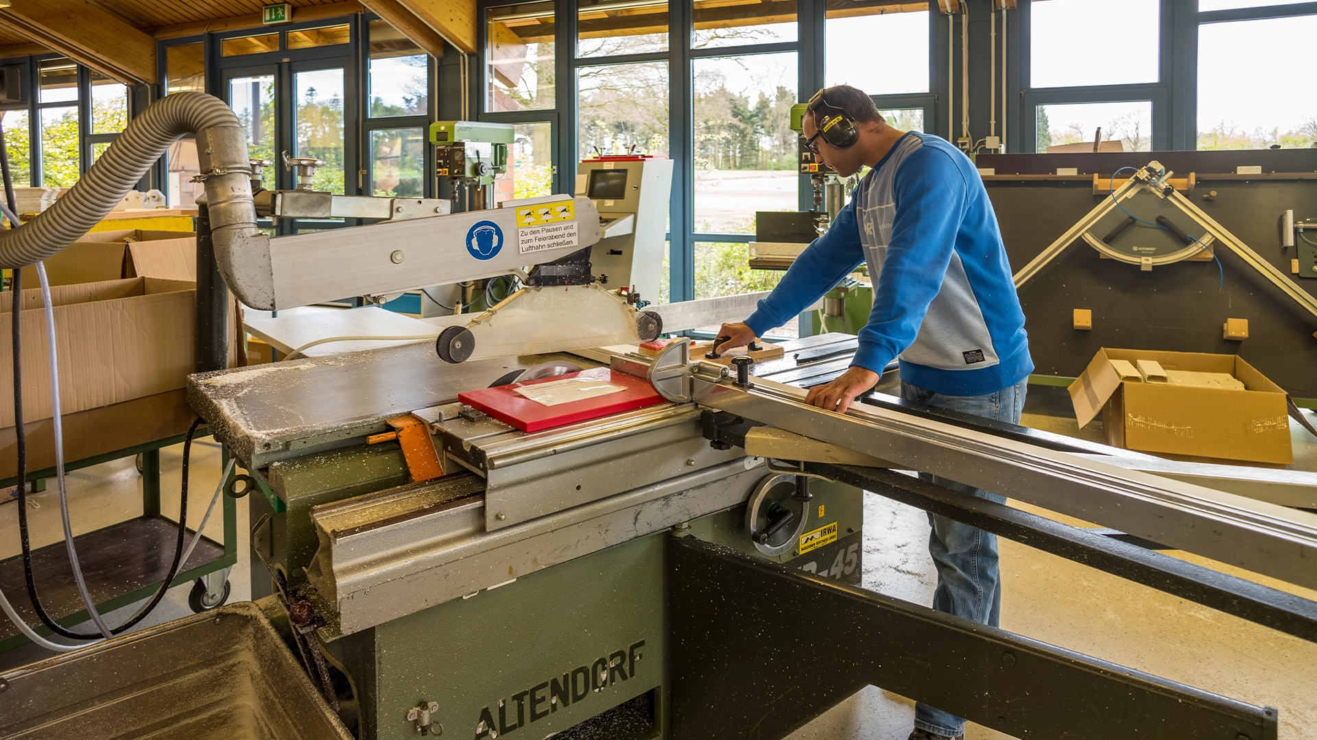 Ein Mann in blauer Jacke und Gehörschutz bedient eine große Holzschneidemaschine in einer Werkstatt mit Fenstern im Hintergrund. Auf dem Tisch liegt ein roter Ordner. Im Hintergrund stehen Kartons und Holzrahmen.