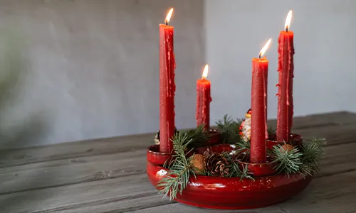Ein Adventskranz mit vier brennenden roten Kerzen auf einem Holztisch. Der Kranz ist mit grünen Tannenzweigen und kleinen Zapfen dekoriert. Der Hintergrund ist neutral und schlicht. Bild verlinkt zum Produkt.