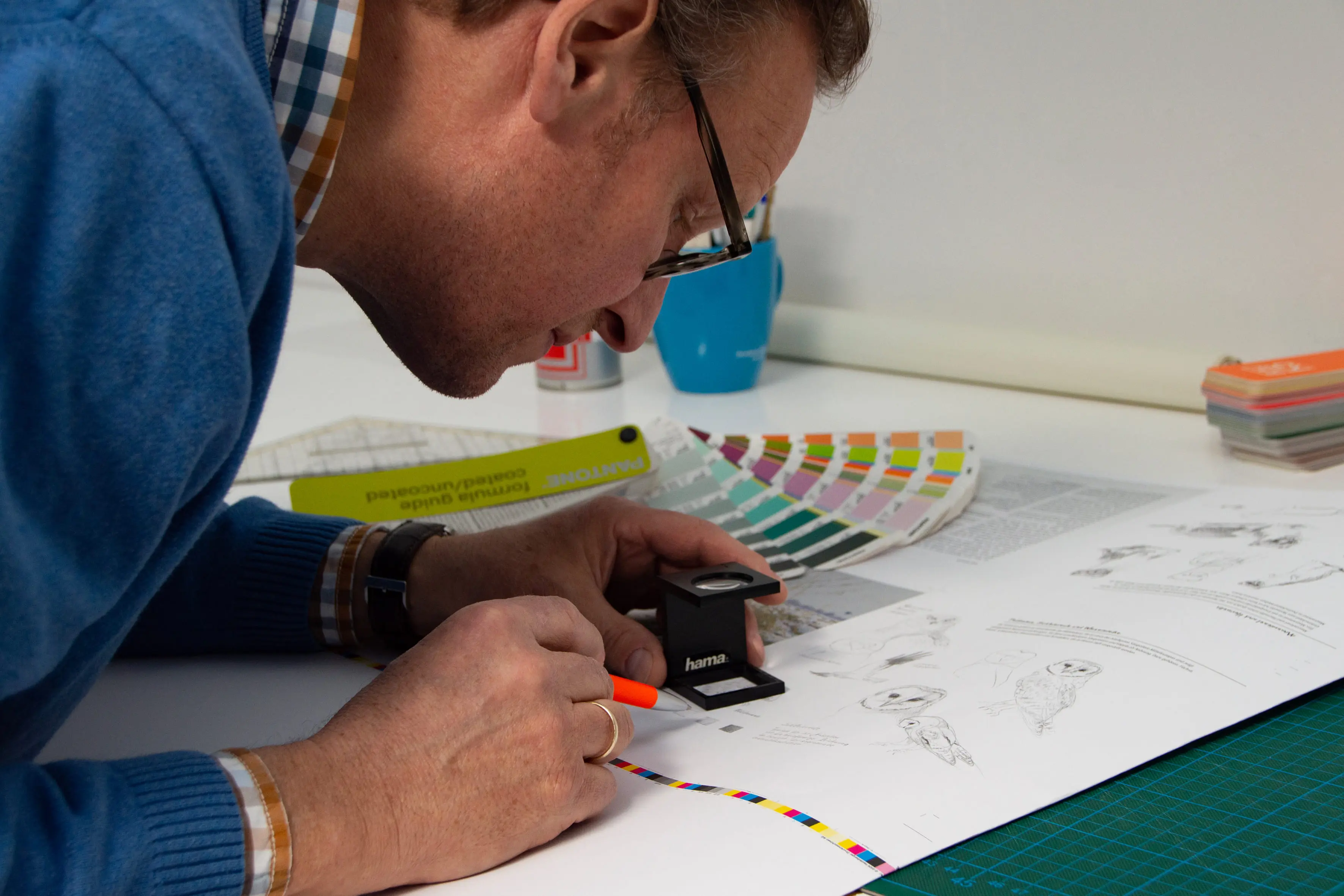 Ein Mann betrachtet mit einer Lupe einen großformatigen Druckbogen auf einem Schreibtisch. Er überprüft farbige Teststreifen und Druckdetails. Neben ihm liegen Farbfächer, Lineale und ein Cutter. Im Hintergrund stehen ein blauer Becher und ein Farbfächers