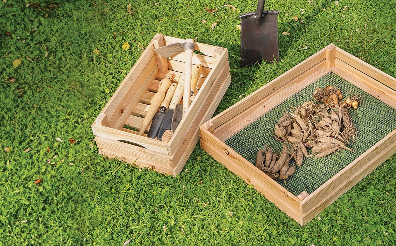 Holzkisten auf Rasen; eine enthält Gartengeräte wie Hacken und Spaten, die andere trockene Blätter und Zwiebeln. Ein Spaten lehnt im Hintergrund an der Kiste. Bild verlinkt zum Produkt.