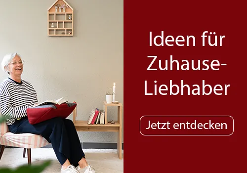 Eine ältere Frau mit grauem Haar sitzt in einem gestreiften Sessel und liest ein Buch, das auf einem roten Buchkissen liegt. Neben ihr steht ein Holztisch mit einem brennenden Kerzenlicht und mehreren Büchern. Bild verlinkt zur Landingpage.