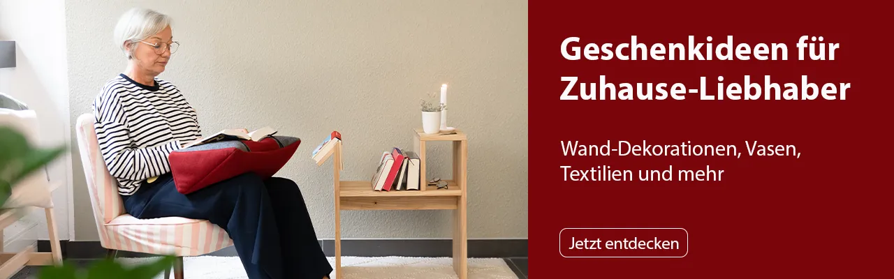 Eine ältere Frau mit grauem Haar sitzt in einem gestreiften Sessel und liest ein Buch, das auf einem roten Buchkissen liegt. Neben ihr steht ein Holztisch mit einem brennenden Kerzenlicht und mehreren Büchern. Bild verlinkt zur Landingpage.