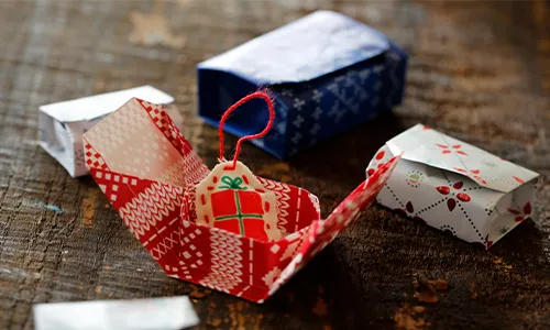 Mehrere kleine, gefaltete Schachteln aus bedrucktem Papier liegen auf einem Holztisch. Eine Schachtel ist offen und zeigt ein Geschenk-Etikett mit rotem Band. Die verbleibenden Schachteln sind geschlossen, Bild verlinkt zum Produkt.