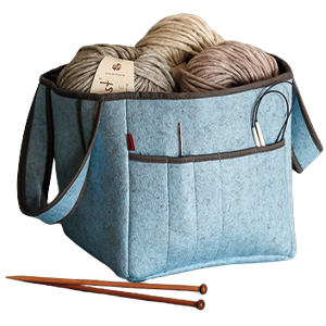 Blaue Filztasche mit mehreren Fächern, gefüllt mit beigen und braunen Wollknäueln, Stricknadeln und einer Rundstricknadel. Zwei Holzstricknadeln liegen davor auf einer rosa Fläche. Bild verlinkt zum Produkt.
