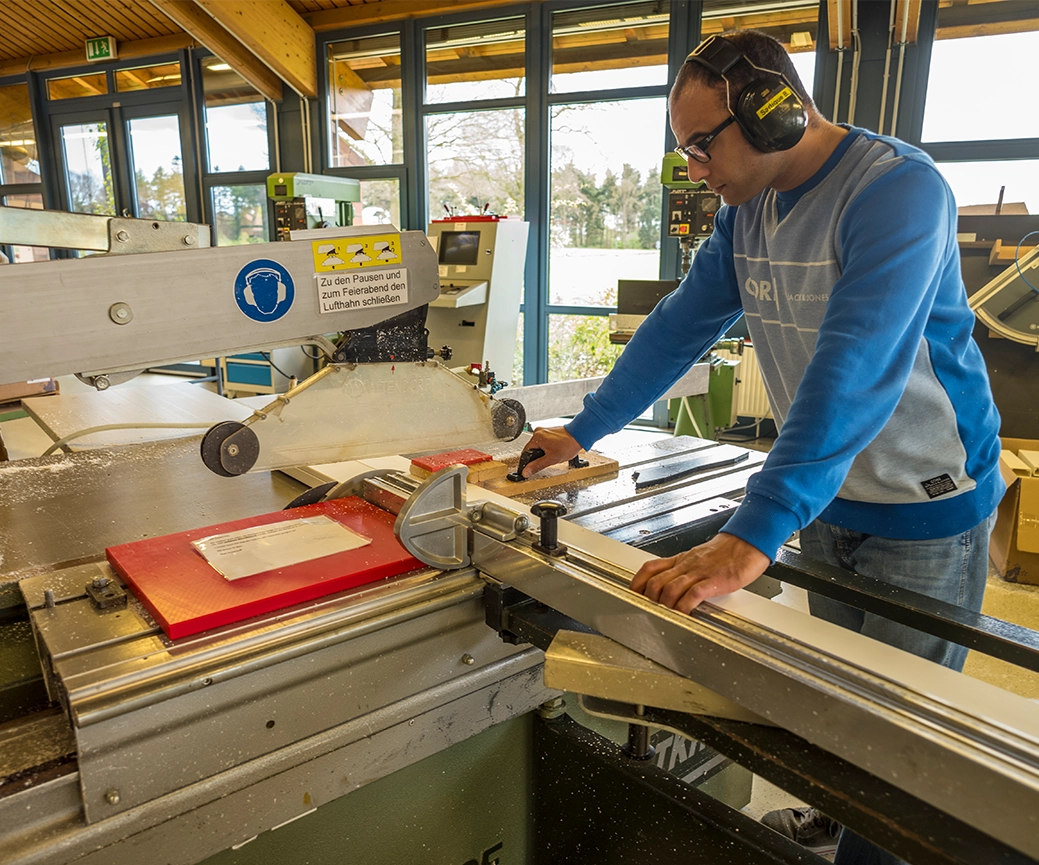 Ein Mann trägt Gehörschutz und arbeitet in einer Holzwerkstatt. Er bedient eine große Säge, um ein Holzstück zu schneiden. Der Raum hat große Fenster, durch die Tageslicht einfällt. Bild verlinkt zum Herstellerportrait. 