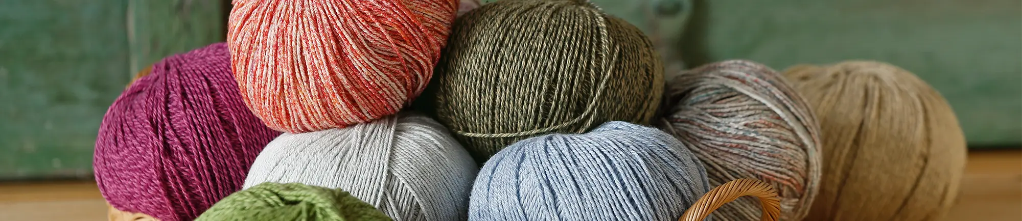 Mehrere Wollknäuel in verschiedenen Farben, darunter Rot, Grün, Grau, Blau und Braun, sind auf einem Tisch arrangiert. Sie liegen teilweise in einem Korb vor einem grünen, verschwommenen Hintergrund. 