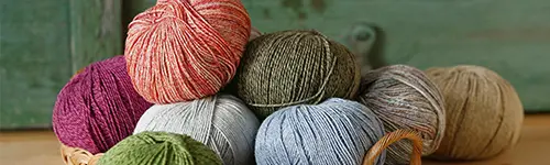 Mehrere Wollknäuel in verschiedenen Farben, darunter Rot, Grün, Grau, Blau und Braun, sind auf einem Tisch arrangiert. Sie liegen teilweise in einem Korb vor einem grünen, verschwommenen Hintergrund. 