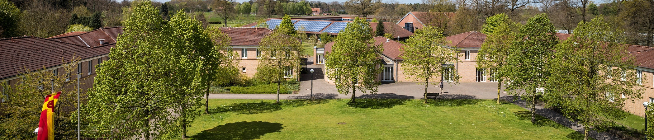 Ein Hof mit roten Ziegeldächern zwischen grünen Bäumen. Vordergrund zeigt eine Rasenfläche, links weht eine rot-gelbe Fahne. Im Hintergrund sind Solarpaneele auf einem Dach sowie mehr Bäume sichtbar.