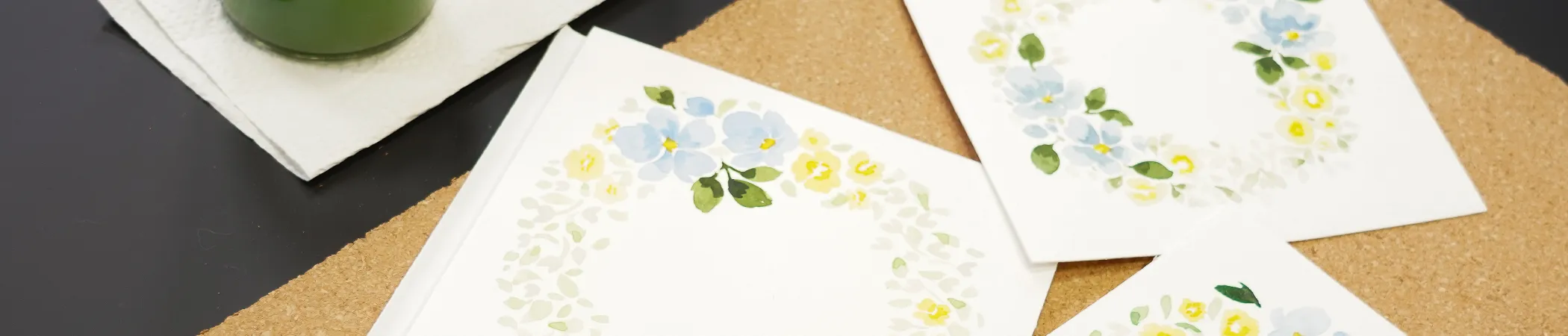 Drei weiße Karten mit floralen Aquarellkränzen in Blau und Gelb liegen auf einer Korkunterlage. Links oben ist ein grünes Glas mit einer weißen Serviette darunter. Die Korkunterlage liegt auf einer schwarzen Oberfläche.