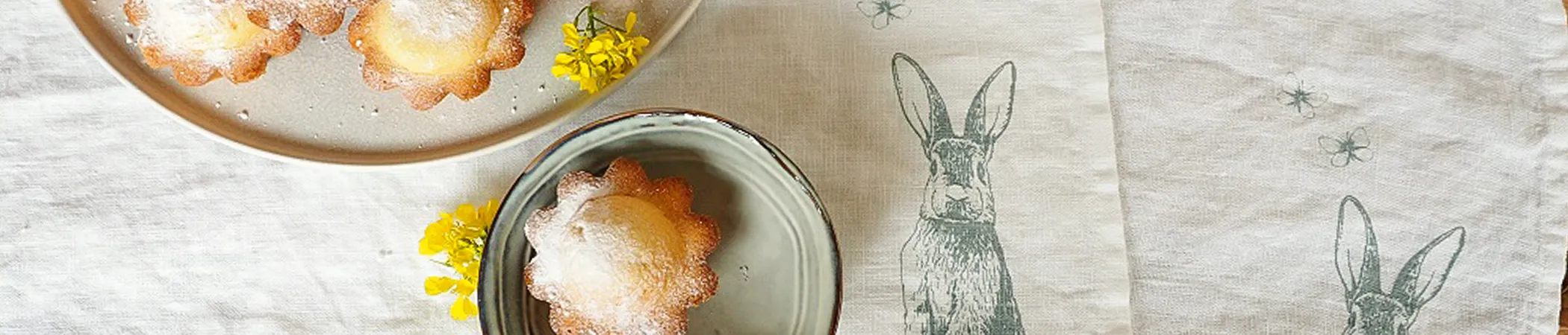 Drei kleine Gebäckstücke auf Tellern, bestäubt mit Puderzucker, liegen auf einem Tischtuch. Rechts im Bild ist ein Häschenmotiv auf das Tuch gedruckt. Neben den Gebäckstücken liegt ein kleiner gelber Blütenzweig.