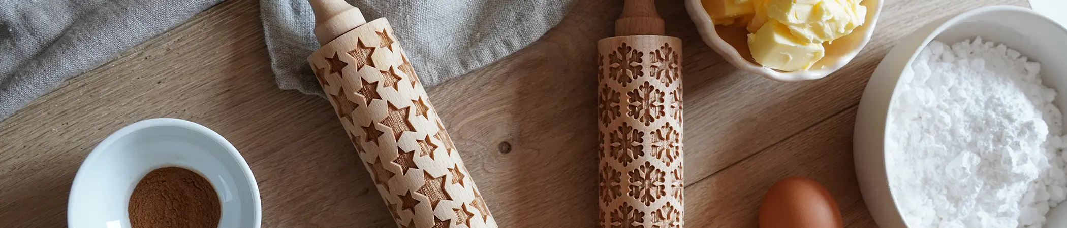 Teigrolle mit Stern- und Schneeflockenmuster, daneben ein Ei, Butter in einer Schale, Mehl und eine Schale mit Zimt auf einem Holztisch.