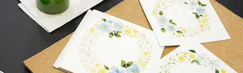 Drei weiße Karten mit floralen Aquarellkränzen in Blau und Gelb liegen auf einer Korkunterlage. Links oben ist ein grünes Glas mit einer weißen Serviette darunter. Die Korkunterlage liegt auf einer schwarzen Oberfläche.