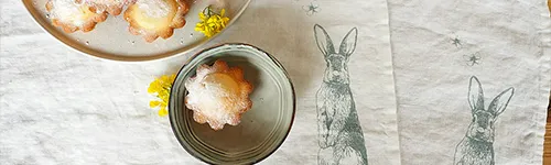 Drei kleine Gebäckstücke auf Tellern, bestäubt mit Puderzucker, liegen auf einem Tischtuch. Rechts im Bild ist ein Häschenmotiv auf das Tuch gedruckt. Neben den Gebäckstücken liegt ein kleiner gelber Blütenzweig.