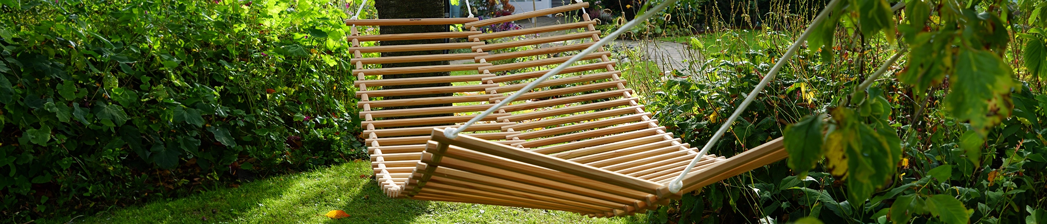 Eine Holz-Hängematte hängt zwischen zwei Bäumen in einem grünen Garten. Die Sonne scheint durch die Blätter und wirft Schatten auf den Rasen. Im Hintergrund sind dichte Büsche und Pflanzen zu sehen. Die Hängematte ist leer und leicht im Wind schwingend.