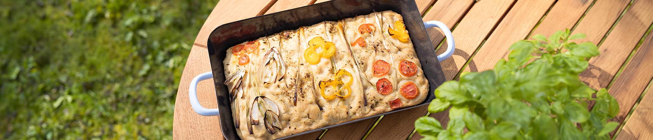 Rechteckige Focaccia in einer schwarzen Auflaufform, garniert mit roten Kirschtomaten, gelben Paprikaringen und Zwiebeln, auf einem Holztisch. Rechts vorne im Bild ist ein grünblättriger Basilikumstrauch zu sehen, im Hintergrund ist eine grüne Wiese.