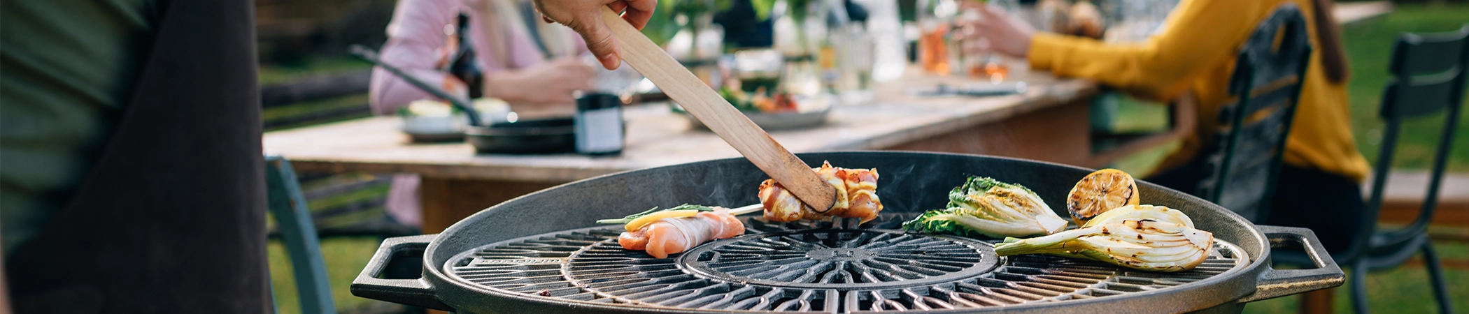 Eine Person grillt auf einem runden Grill. Verschiedene Lebensmittel, darunter Fleisch und Gemüse, liegen auf dem Rost. Im Hintergrund sitzt eine Gruppe von Menschen an einem Holztisch im Freien und isst, trinkt oder unterhält sich.