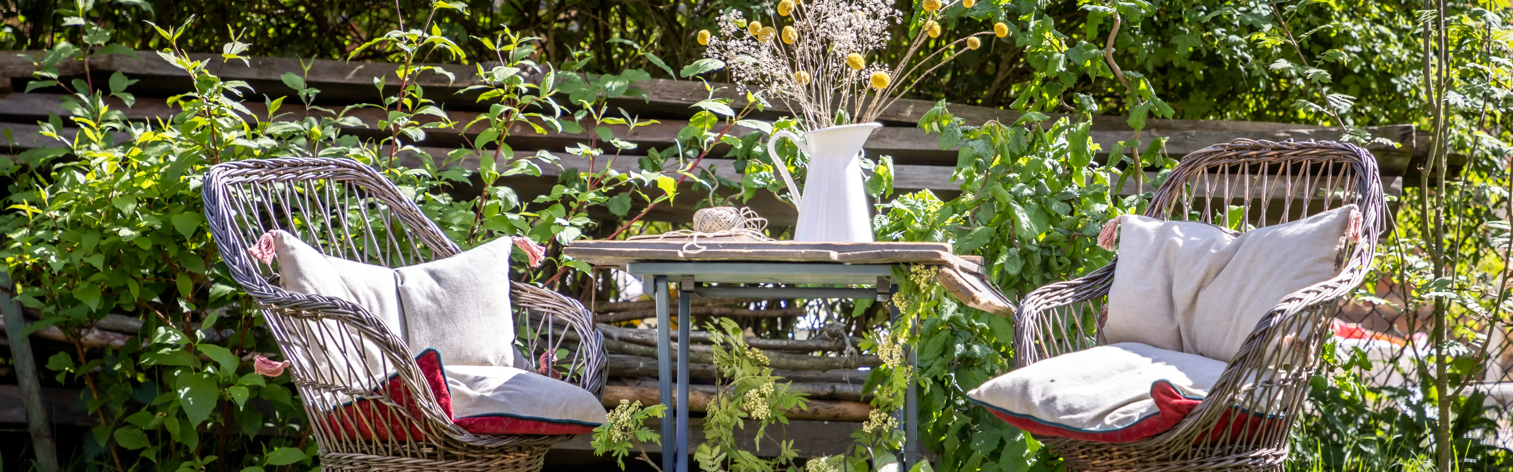 Zwei geflochtene Stühle mit hellen Kissen stehen um einen kleinen Holztisch in einem Garten. Auf dem Tisch befindet sich eine weiße Vase mit Trockenblumen. Hinter der Sitzgruppe sind grüne Pflanzen und ein Holzzaun zu sehen.