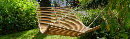 Eine Holz-Hängematte hängt zwischen zwei Bäumen in einem grünen Garten. Die Sonne scheint durch die Blätter und wirft Schatten auf den Rasen. Im Hintergrund sind dichte Büsche und Pflanzen zu sehen. Die Hängematte ist leer und leicht im Wind schwingend.