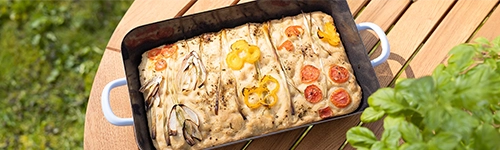 Rechteckige Focaccia in einer schwarzen Auflaufform, garniert mit roten Kirschtomaten, gelben Paprikaringen und Zwiebeln, auf einem Holztisch. Rechts vorne im Bild ist ein grünblättriger Basilikumstrauch zu sehen, im Hintergrund ist eine grüne Wiese.