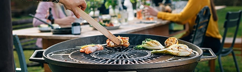Eine Person grillt auf einem runden Grill. Verschiedene Lebensmittel, darunter Fleisch und Gemüse, liegen auf dem Rost. Im Hintergrund sitzt eine Gruppe von Menschen an einem Holztisch im Freien und isst, trinkt oder unterhält sich.