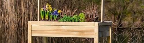 Hölzerner Blumenkasten auf Beinen, bepflanzt mit gelben, lila und blauen Blumen sowie grünen und braunen Pflanzen. Im Hintergrund unscharf Bäume und ein Teich.