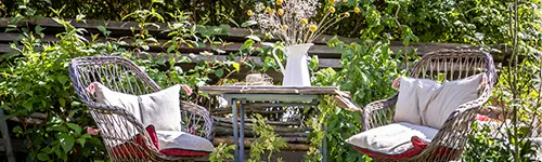 Zwei geflochtene Stühle mit hellen Kissen stehen um einen kleinen Holztisch in einem Garten. Auf dem Tisch befindet sich eine weiße Vase mit Trockenblumen. Hinter der Sitzgruppe sind grüne Pflanzen und ein Holzzaun zu sehen.