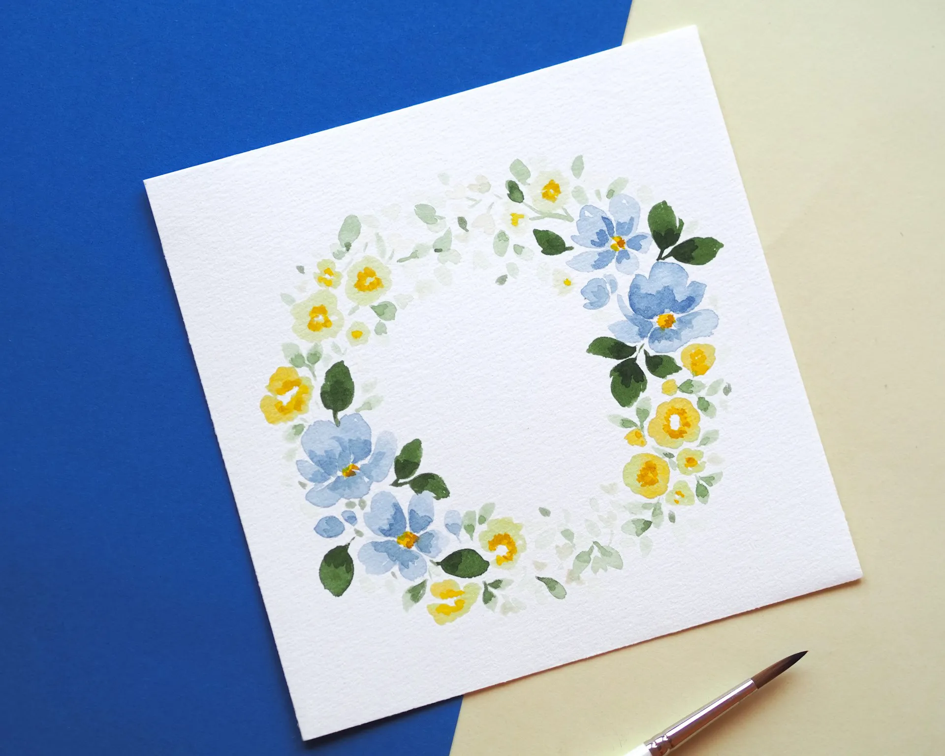 Aquarellgemälde eines Kranzes aus blauen und gelben Blumen mit grünen Blättern, auf weißem Papier. Hintergrund in Blau und Beige, unten rechts ein Pinsel.