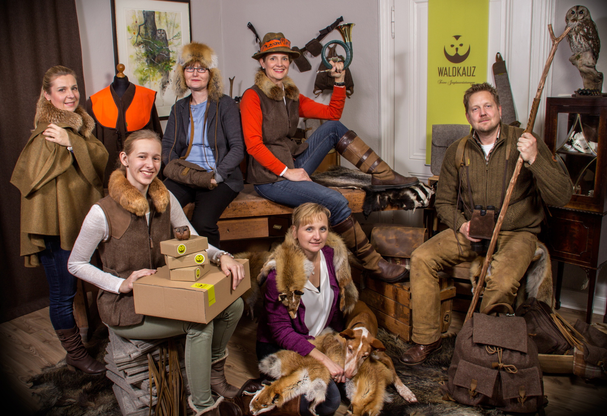 Gruppe von sechs Personen in Jagdkleidung posieren in einem Raum mit Holzmöbeln und Jagdutensilien. Eine Person hält Kartons mit Wildroar-Logo, eine sitzt mit einem ausgestopften Tier, ein Hund liegt vorne.