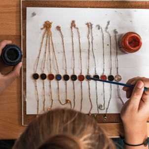 Frau Schnieke sitzt an einem Holztisch und malt ein Blatt Papier mit Linien und Punkten in verschiedenen Farben. Es hält einen Pinsel in der rechten Hand und hat zwei Farbtöpfe, einen schwarzen und einen roten, auf dem Tisch stehen. 