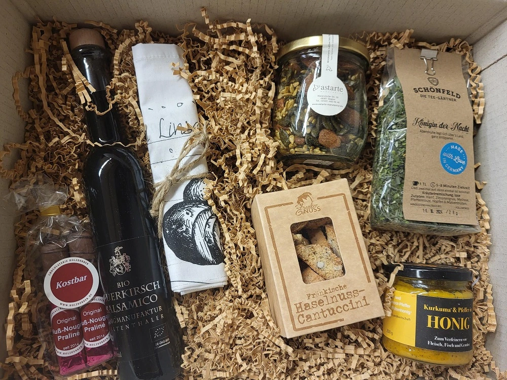 Eine Geschenkbox gefüllt mit Produkten: Flasche Bio-Schwarzkirsche-Balsamico, zwei Packungen Pralinen, bedrucktes Tuch, Glas mit Nüssen, Packung getrocknete Kräuter, Packung Haselnuss-Cantuccini, Glas Honig. Verpackt in Papier.Bild verlinkt zur Winterbox.