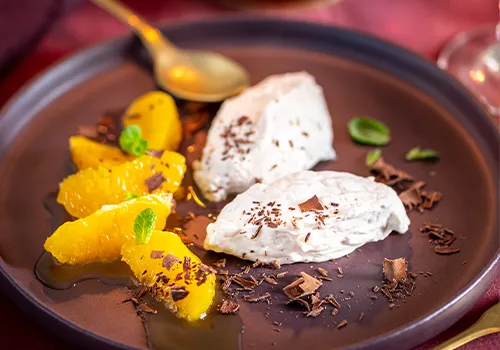 Auf dunkelviolettem Teller liegen zwei Portionen Sahnedessert, garniert mit Schokoladenraspeln und Minzblättern. Daneben sind Orangenscheiben in Sirup angeordnet. Ein goldener Löffel liegt am Tellerrand. Bild verlinkt zum Rezept