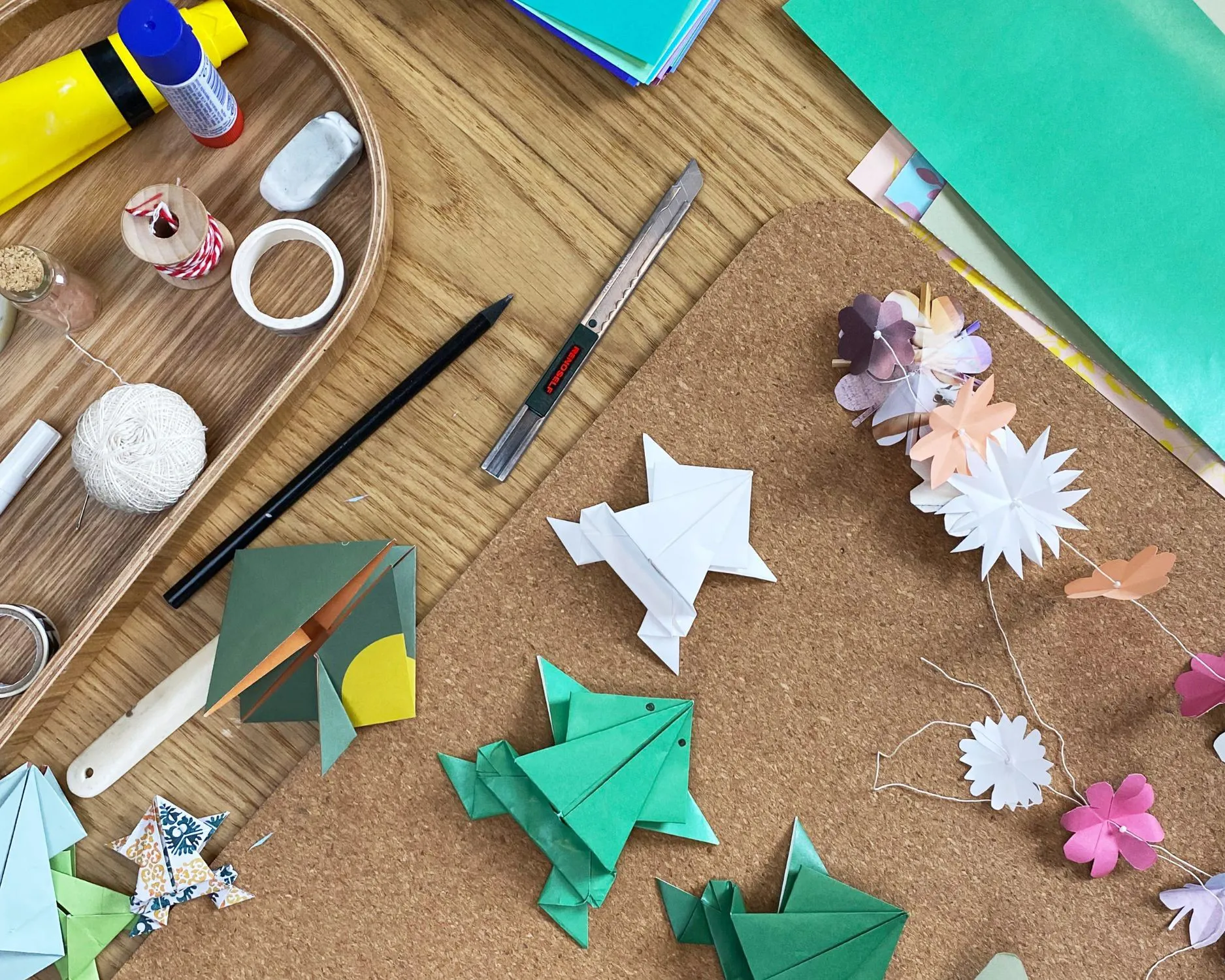 Verschiedene Origami-Figuren, darunter Frösche und Blumen, liegen auf einem Korkbrett und einem Tisch. Daneben befinden sich Bastelutensilien wie Schere, Kleber und Faden. Bild verlinkt zum Workshop.