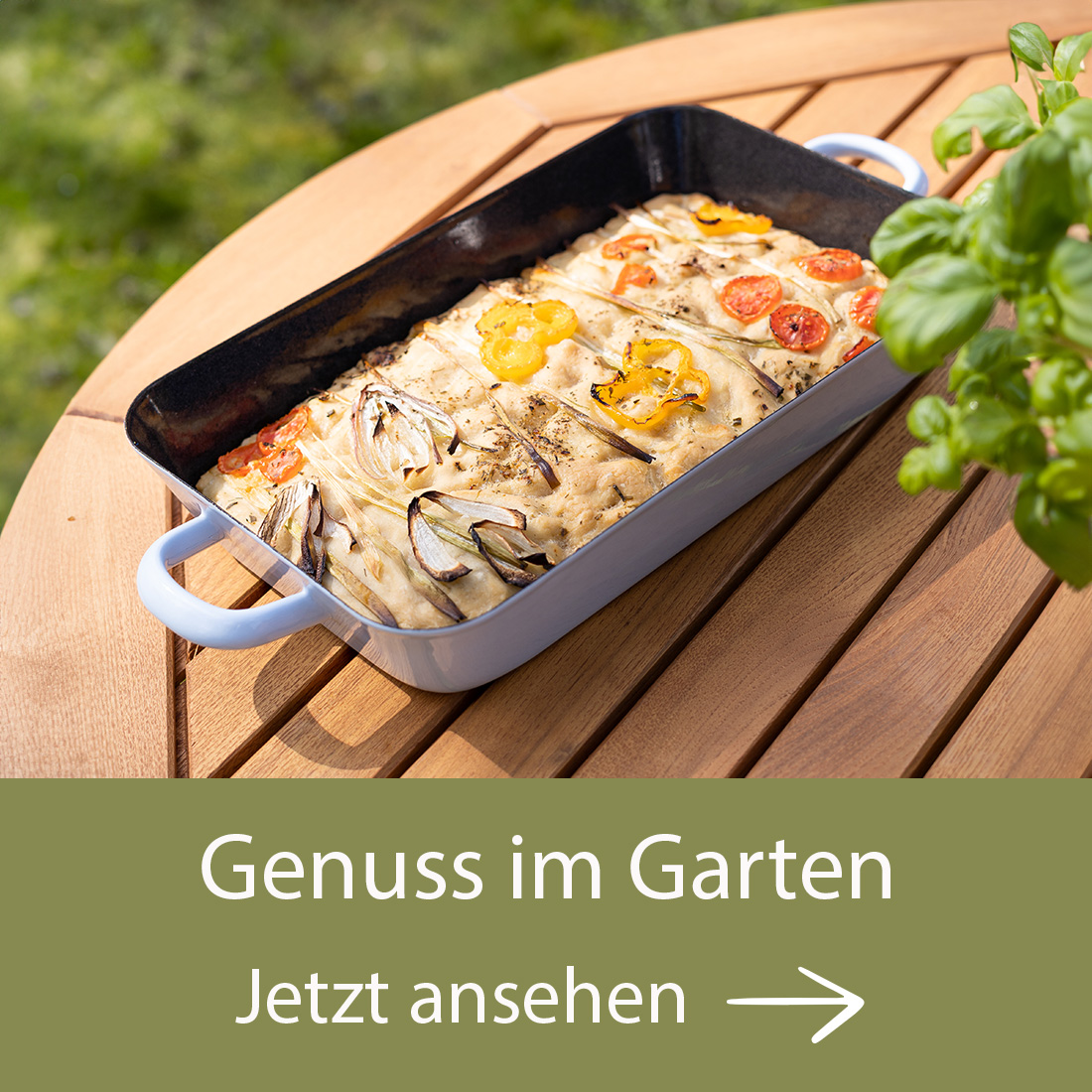 Eine weiße Auflaufform mit gebackenem Gericht, garniert mit gelben und roten Tomatenscheiben, steht auf einem Holztisch im Freien. Daneben ist ein Basilikumtopf zu sehen. Bild verlinkt auf die Landingpage Genuss im Garten.