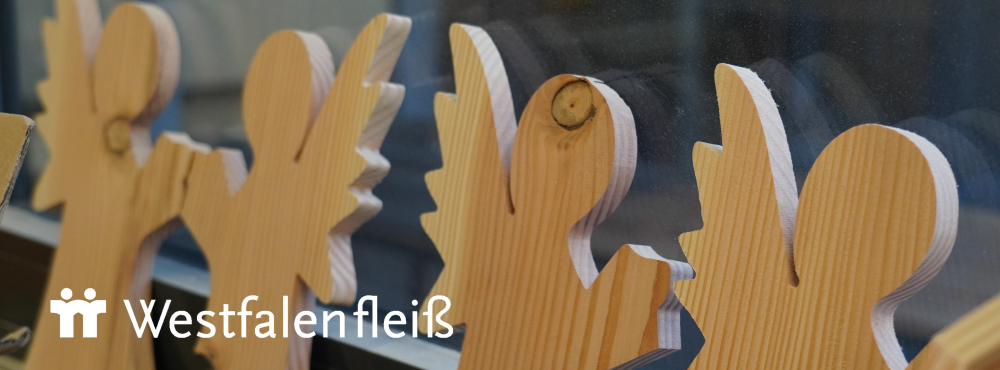 Holzfiguren vor einem Fenster; darunter befindet sich das Logo von Westfalenfleiß mit stilisierten Figuren und dem Schriftzug Westfalenfleiß.