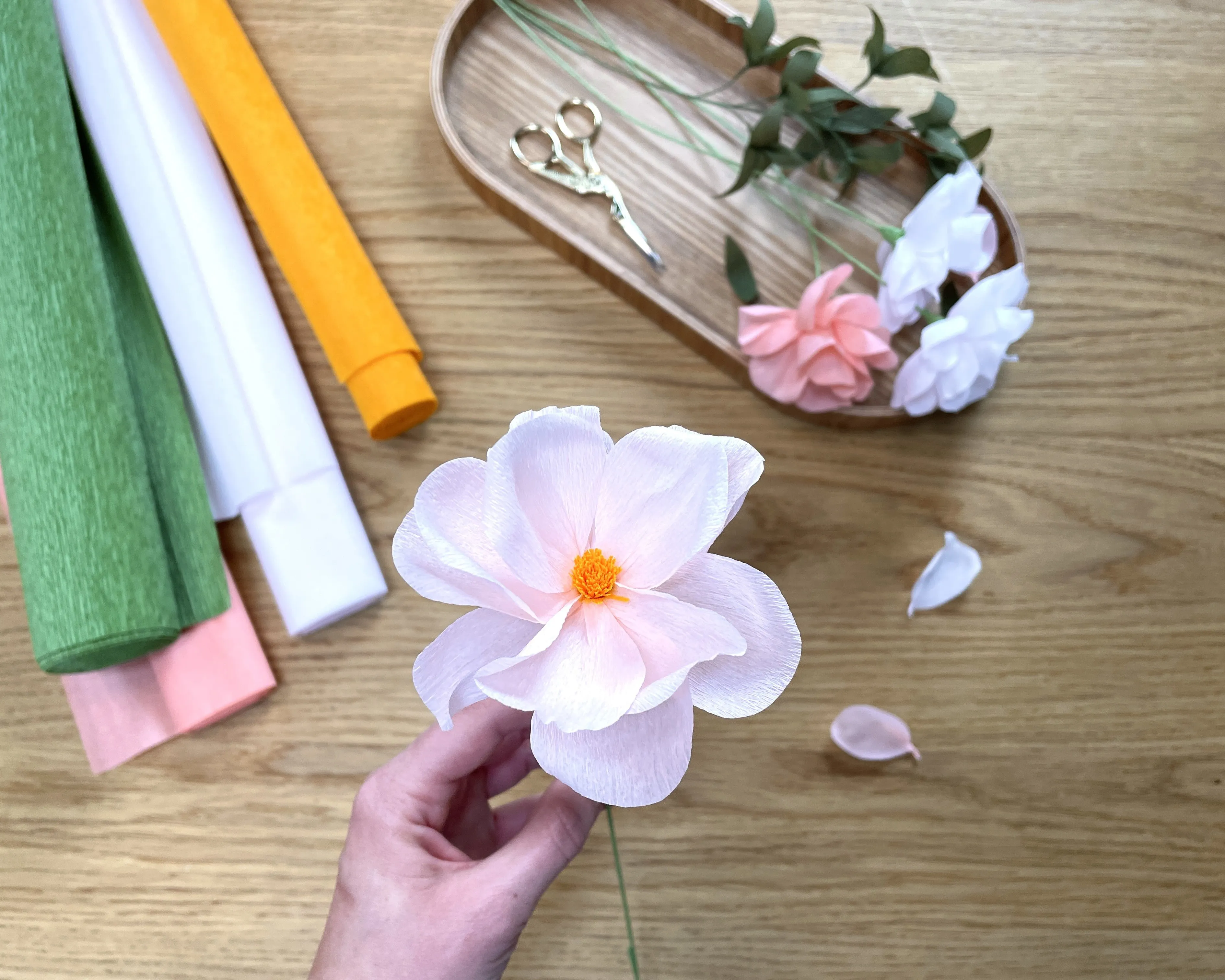 Hand hält eine rosa Papierblume mit gelber Mitte über einem Holztisch. Im Hintergrund liegen bunte Krepppapierrollen, eine Schere und weitere Papierblumen auf einem ovalen Holztablett. Einige Papierblätter sind verteilt. Bild verlinkt zum Workshop.